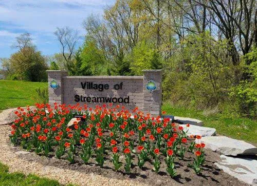 Brick sign reading “Village of Streamwood” behind a bed of red tulips, surrounded by green grass and trees under a partly cloudy sky.