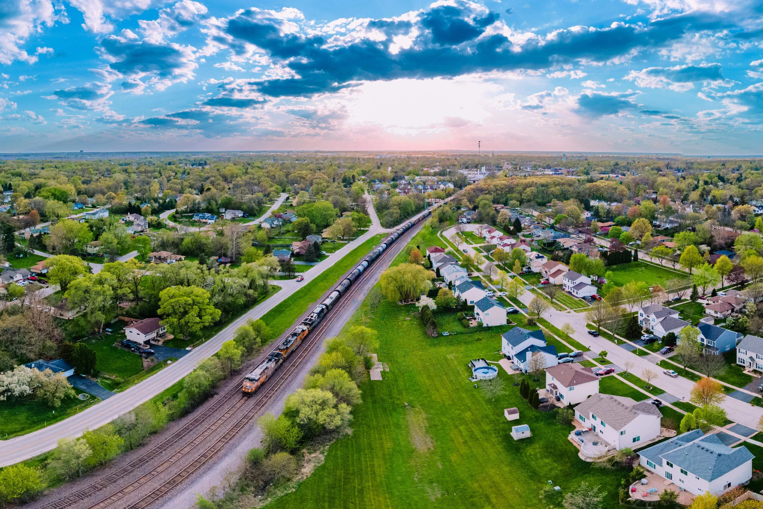 Aerial view of a residential neighborhood with a train traveling along railroad tracks surrounded by houses, trees, and greenery under a partly cloudy sky at sunset.