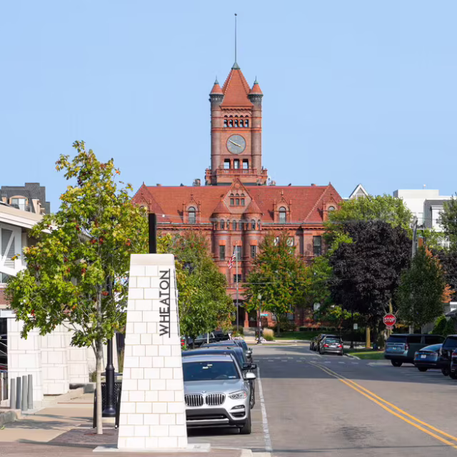 A red-brick courthouse with a clock tower stands at the end of a street lined with parked cars, trees, and a sign reading “Wheaton.”.