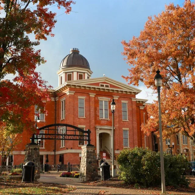 Red-brick courthouse with dome roof, arched entry, and iron gate, surrounded by autumn trees with orange and red leaves under a blue sky.