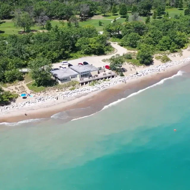 Aerial view of a building near a sandy shoreline with turquoise water, rocky barriers, parked cars, and dense green trees in the background.