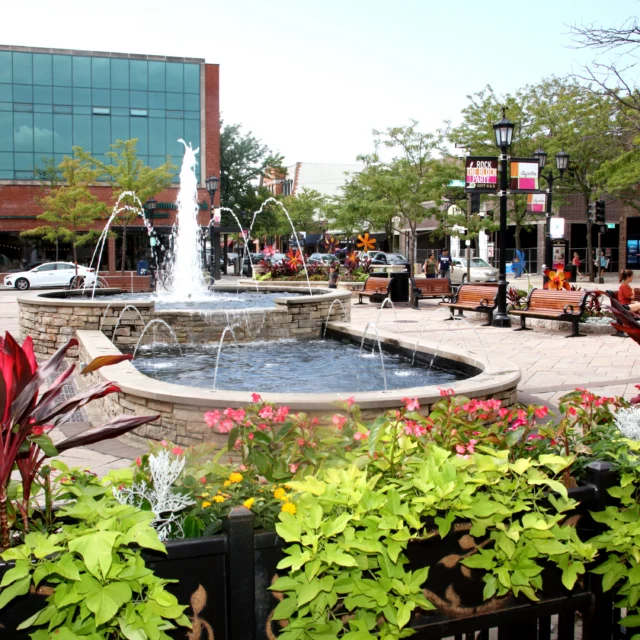 A city plaza with a large fountain, flower beds, benches, and surrounding storefronts on a sunny day.