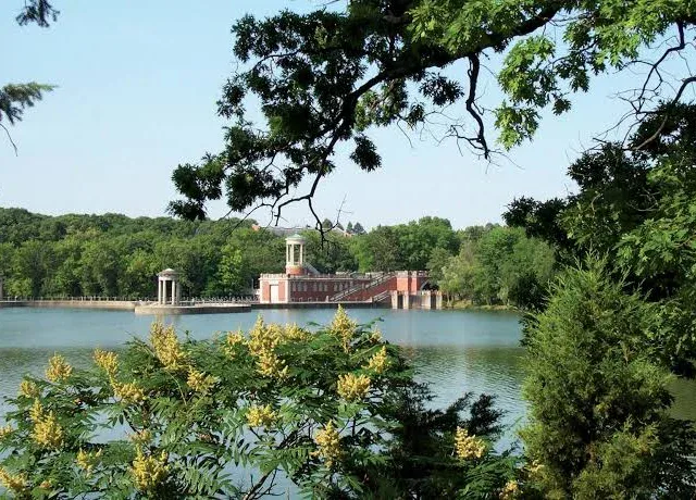 A red brick pavilion with white columns stands by a lake, surrounded by trees and yellow-flowering shrubs under a clear sky.